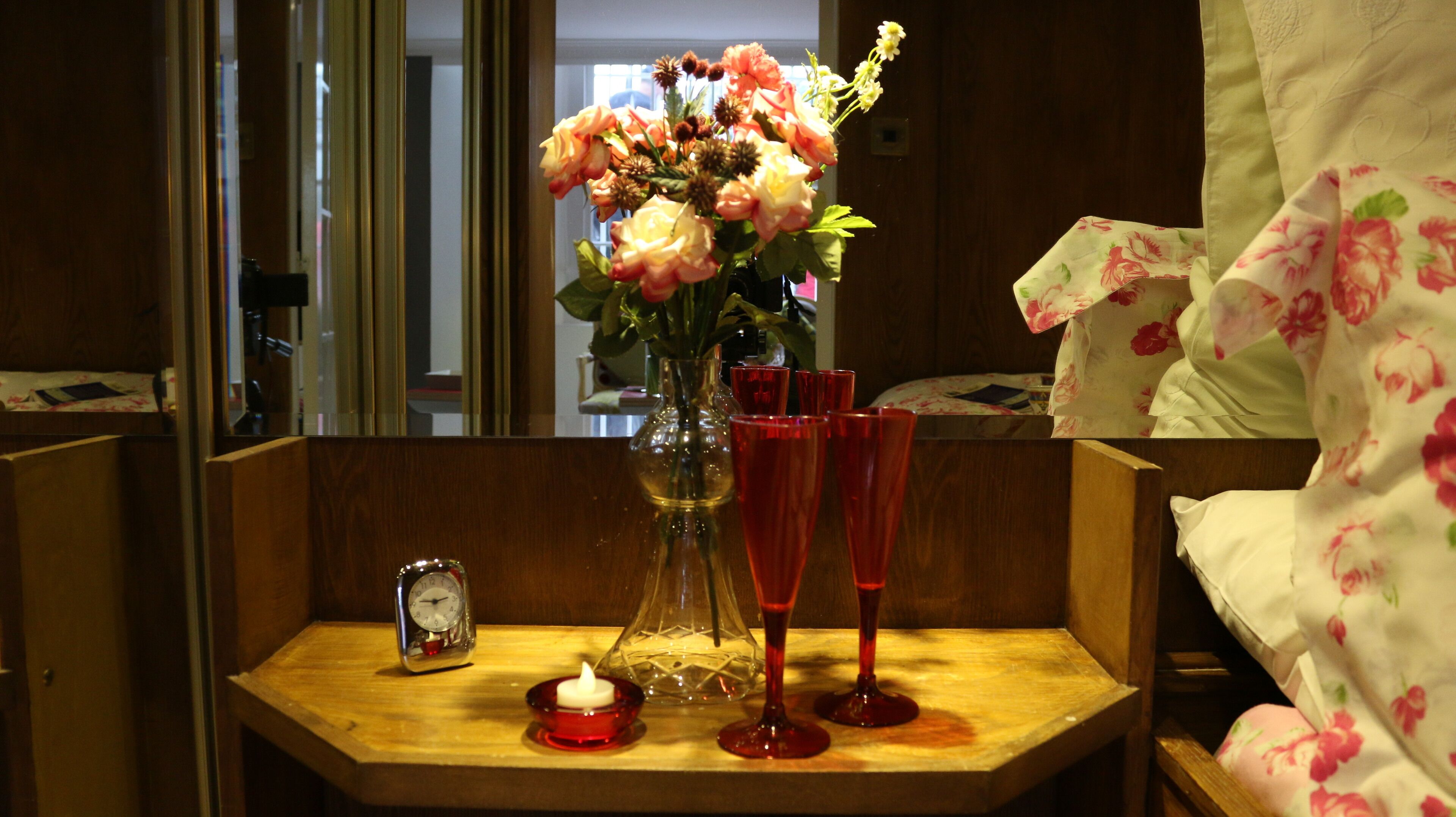 A wooden bedside table with a vase of flowers, two red glasses, a small red candle, and a clock.