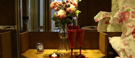 A wooden bedside table with a vase of flowers, two red glasses, a small red candle, and a clock.