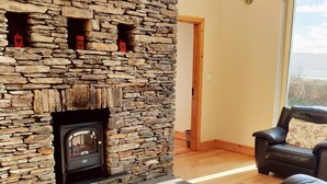 A living room with a stone fireplace, leather sofa, and a view through a large window. - Traditional style cottage free WiFi short walk from Reenroe Beach Ballinskelligs (Ballinskelligs)