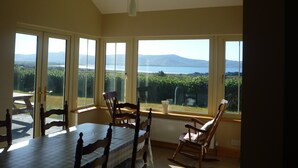 A dining area with a table and chairs, large windows with a view of a lake and mountains, and a hanging light fixture. - Traditional style cottage free WiFi short walk from Reenroe Beach Ballinskelligs (Ballinskelligs)