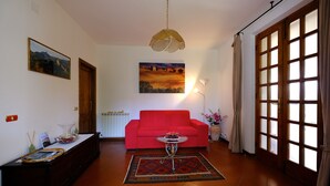 A living room with a red sofa, a coffee table, a painting, and a large window with curtains.