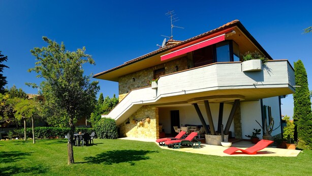 A two-story house with a stone facade, a red awning, and a balcony with red lounge chairs.