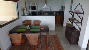 Un comedor con una mesa dispuesta para cuatro personas, rodeada de sillas de mimbre, una cocina al fondo y una estantería decorativa con diversos objetos.