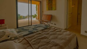 Quarto com cama, cadeira, mesa vermelha e porta de vidro corrediça para uma varanda.