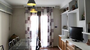 A dining area with a glass table and chairs, a television on a white shelf, and a window with floral curtains.