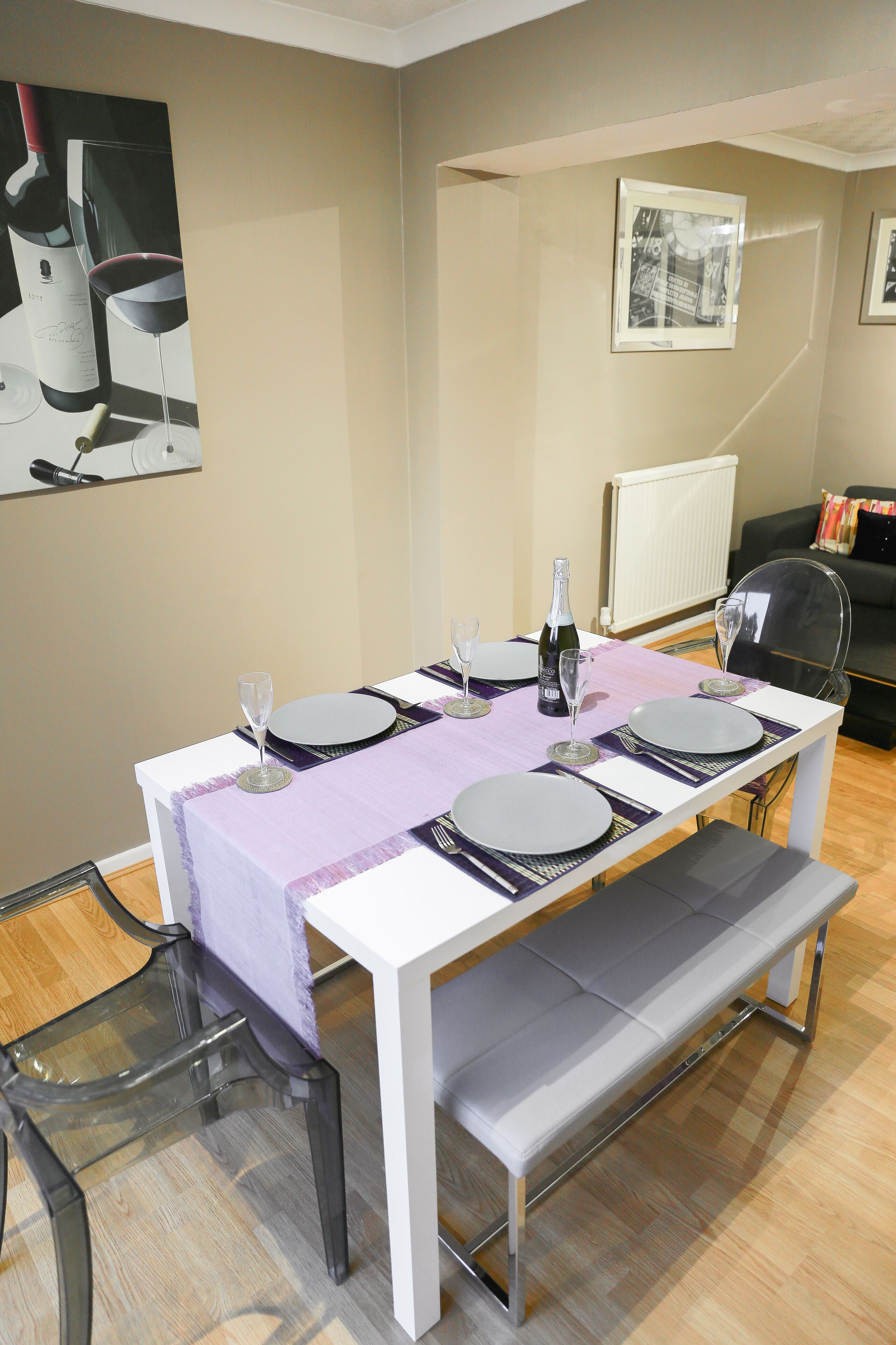 Comedor con una mesa dispuesta para cuatro, incluyendo copas de vino y una botella. Hay sillas con estructura metálica y un banco. La habitación tiene un radiador y un cuadro en la pared.