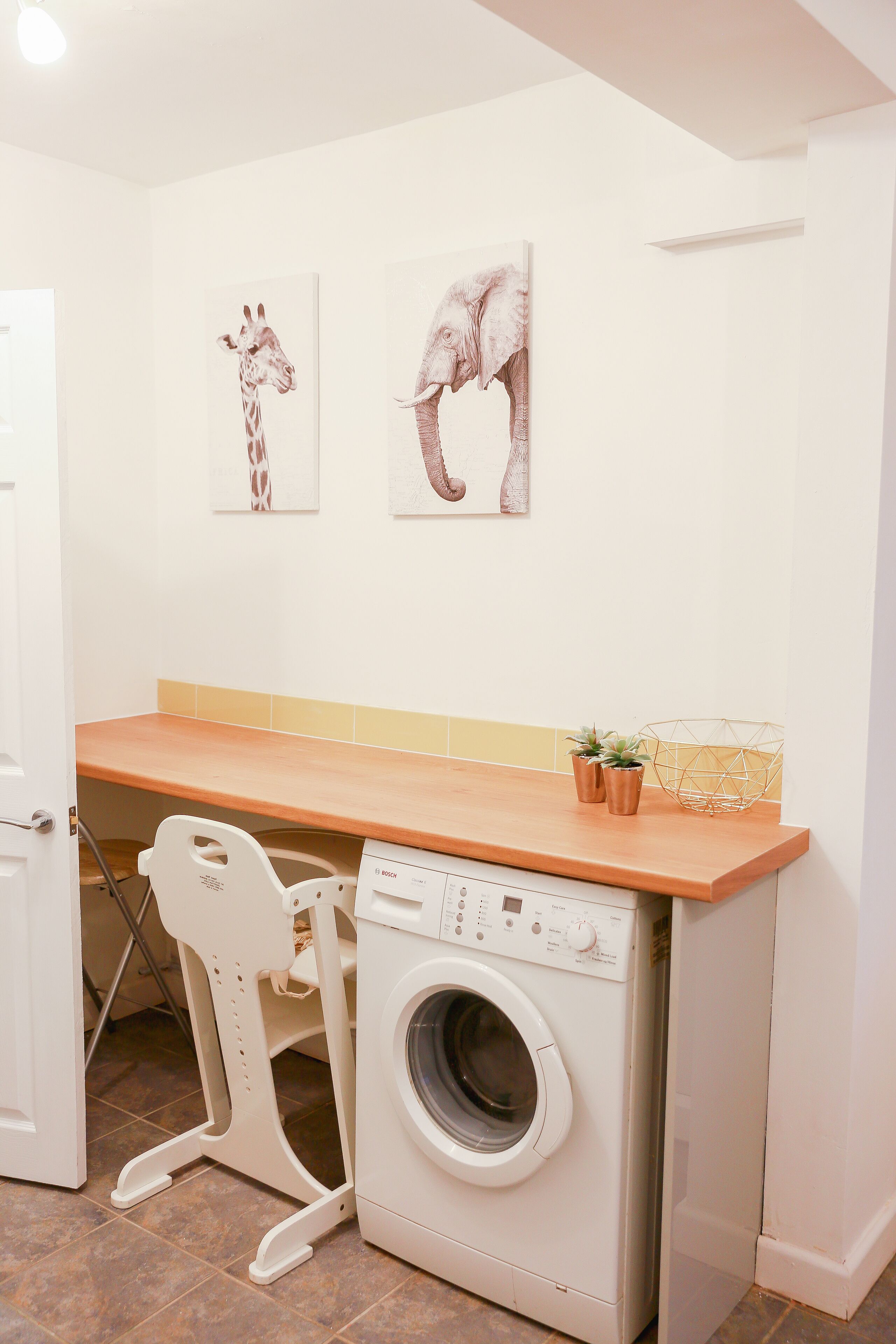 Un espace buanderie compact comprenant un lave-linge, une chaise et deux œuvres d’art fixées au mur, représentant une girafe et un éléphant.