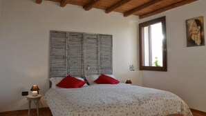 Un dormitorio con techo de madera, una cama con ropa de cama floral, almohadas rojas, una mesita de noche con una lámpara y una ventana con un cuadro.