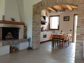 Een ruime keuken met een open haard, een eethoek met tafel en stoelen, en een groot raam voor natuurlijk licht.