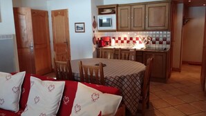 A compact kitchen with wooden cabinets, a microwave, and a tiled backsplash. There is a dining table with chairs and a bed with red and white bedding in the foreground.