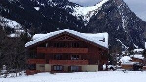 A wooden chalet with a snow-covered roof, multiple balconies, and a mountainous backdrop.