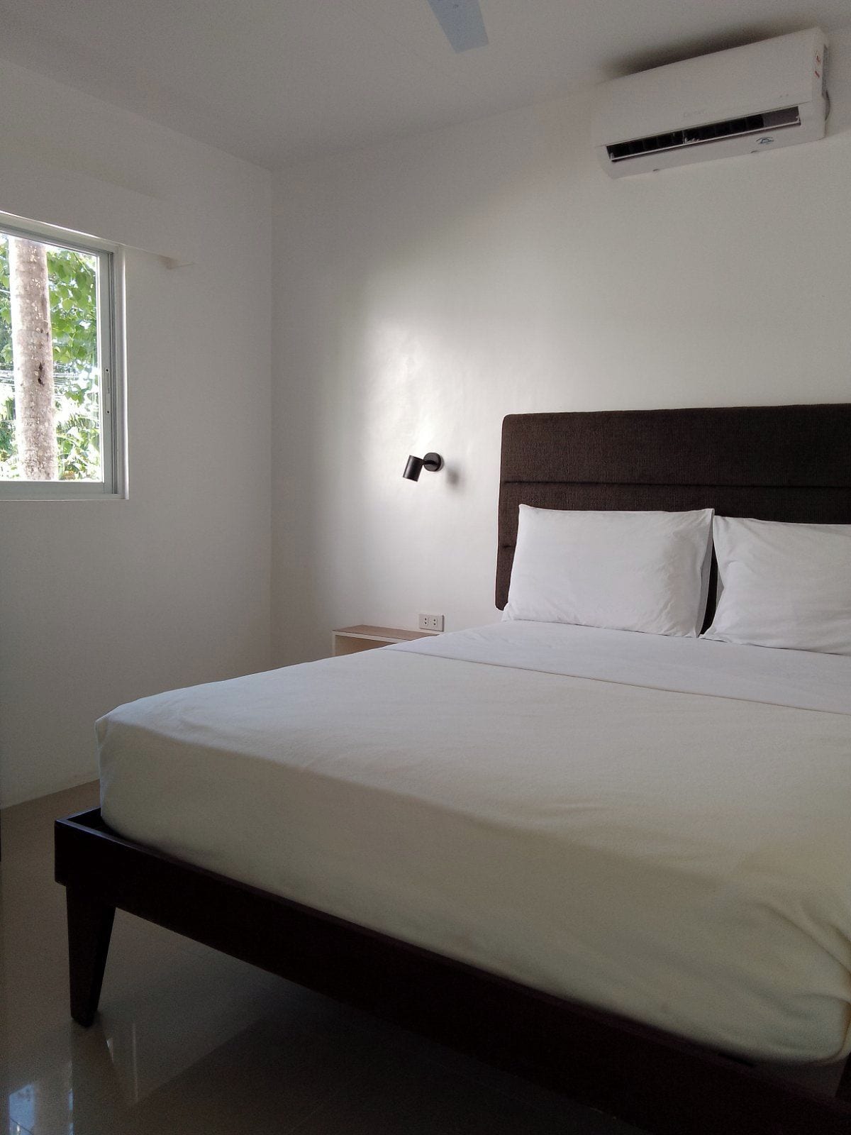 A bedroom with a bed, a nightstand, an air conditioning unit, and a window with a view of trees.