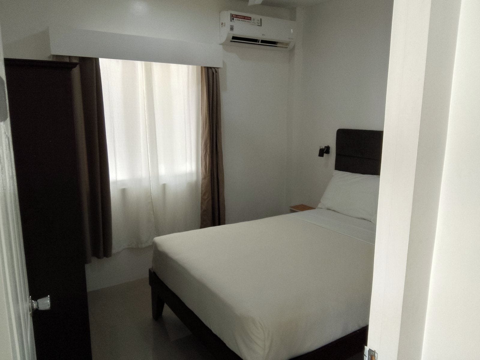 A hotel room with a bed, a nightstand, a wall-mounted air conditioner, and a window with curtains.