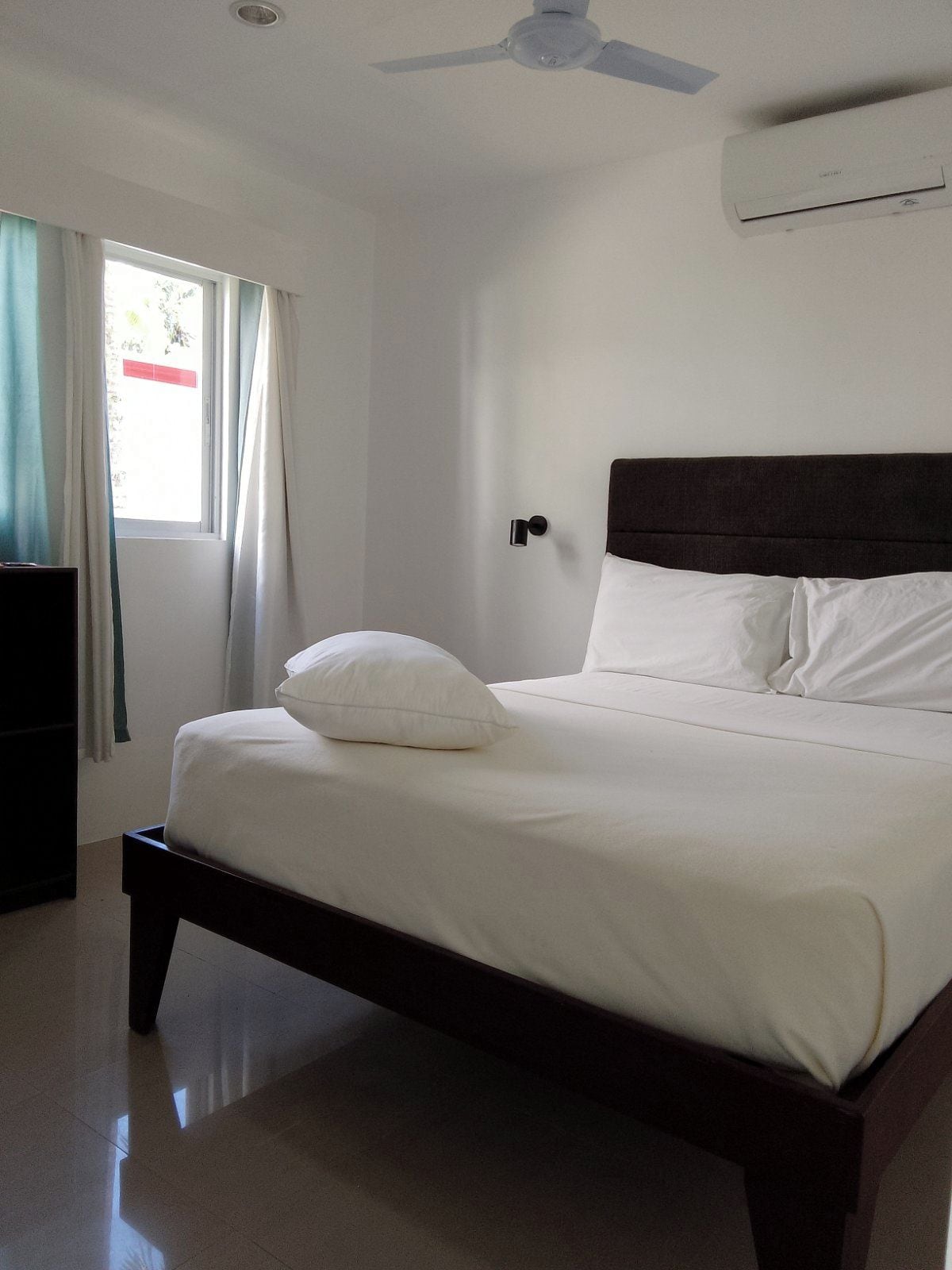 Un dormitorio con cama, ventilador de techo, aire acondicionado y ventana con cortinas.