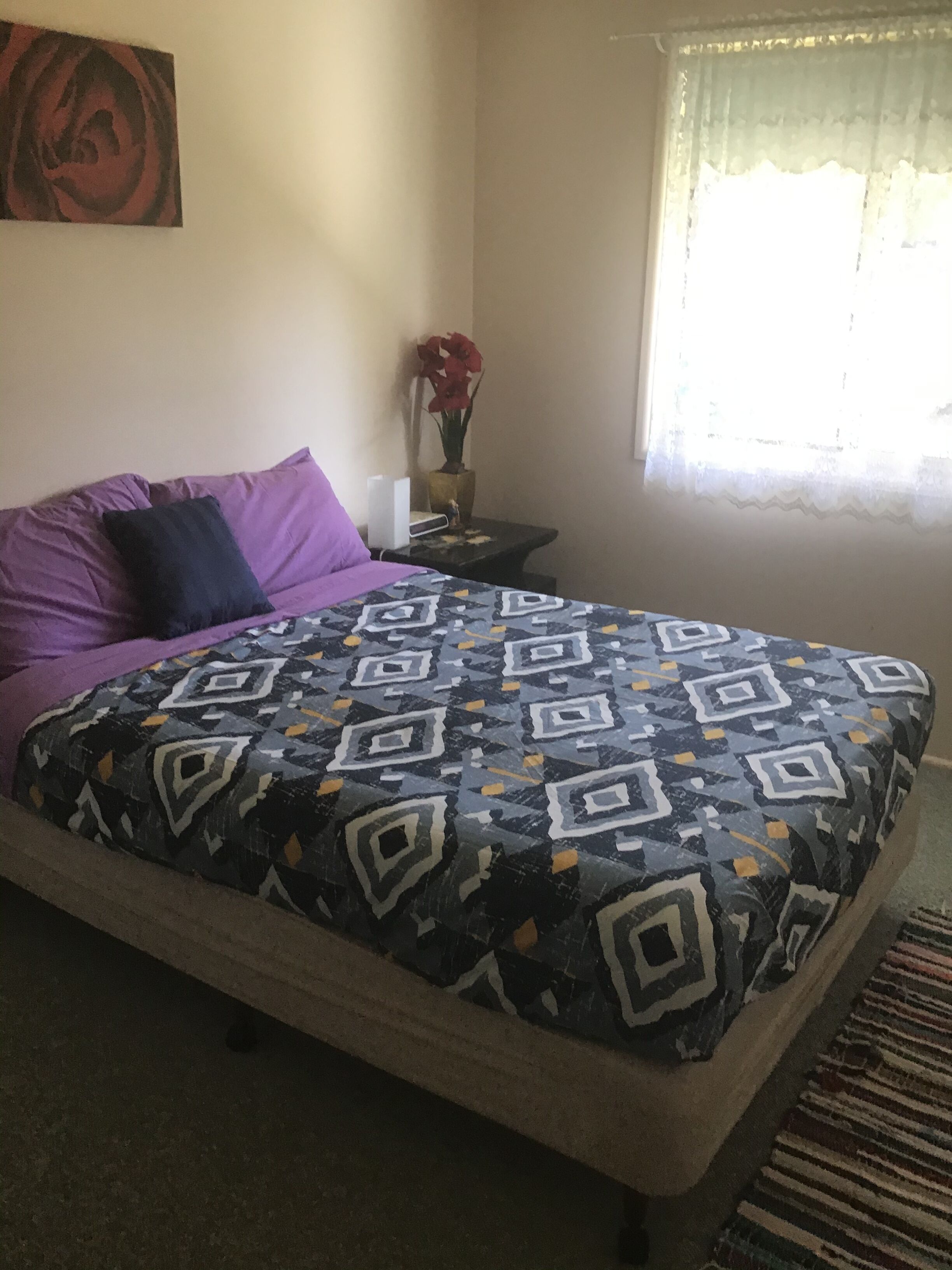 A bedroom with a bed, a nightstand, a vase with flowers, and a window with curtains.