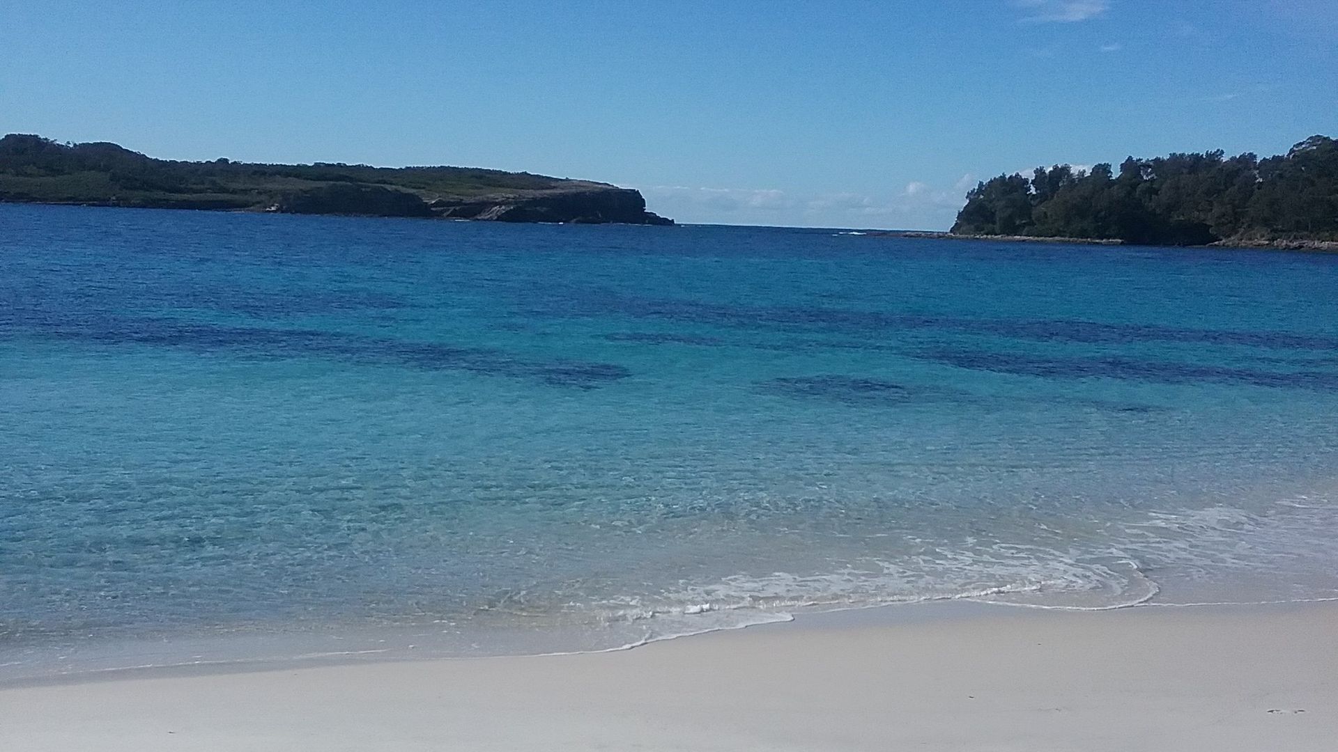 A beach with clear blue water, sandy shore, and distant hills.
