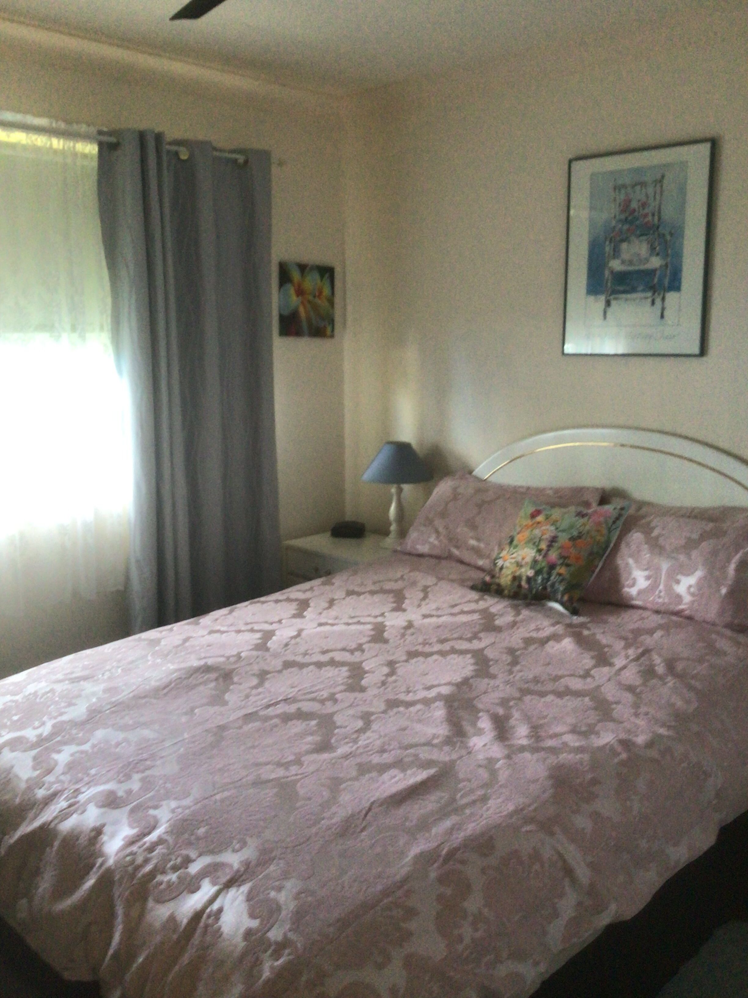 A bedroom with a bed, a nightstand, a lamp, a window with curtains, and a painting on the wall.