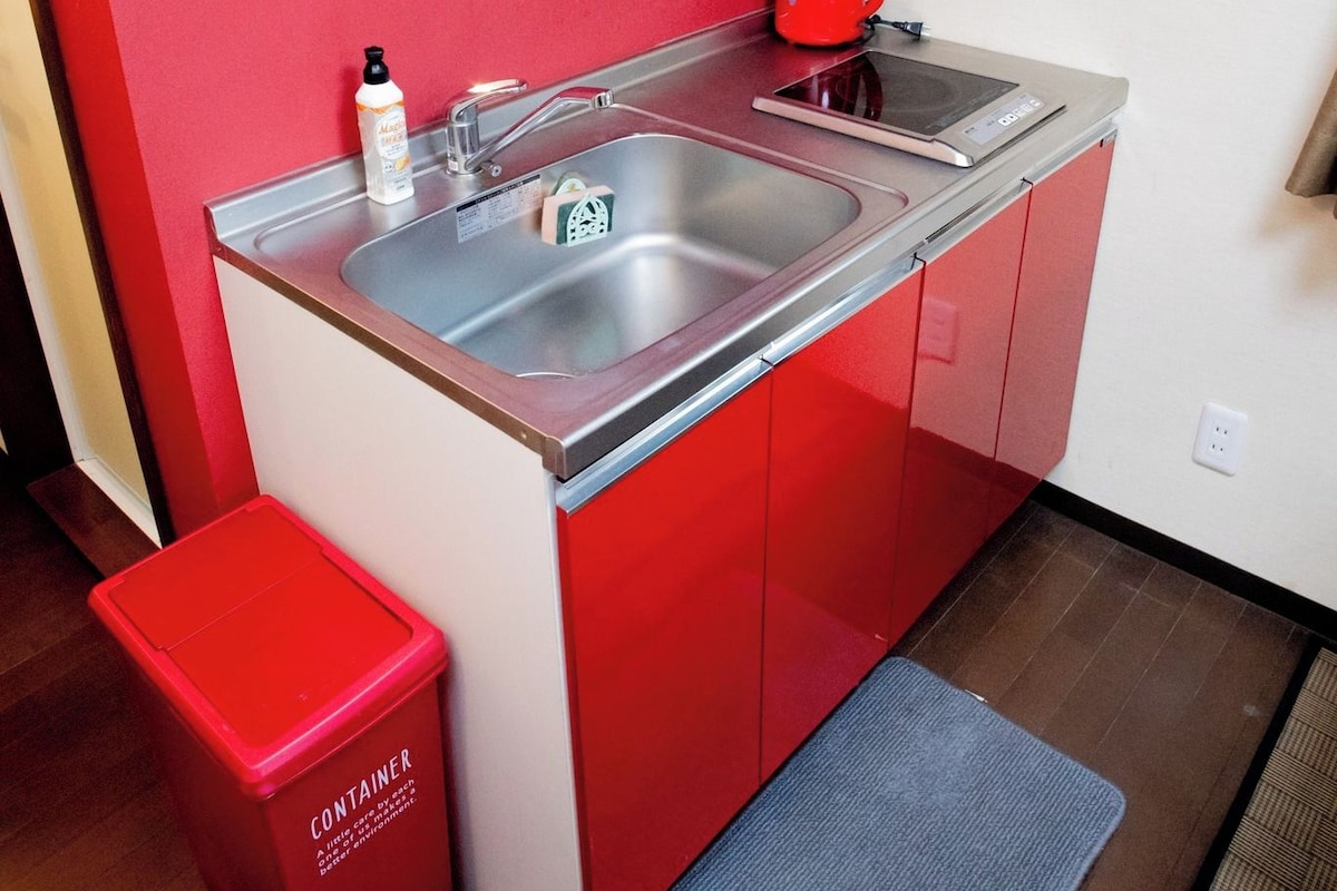 Une petite cuisine équipée avec un évier en acier inoxydable, une bouilloire rouge et une petite poubelle.