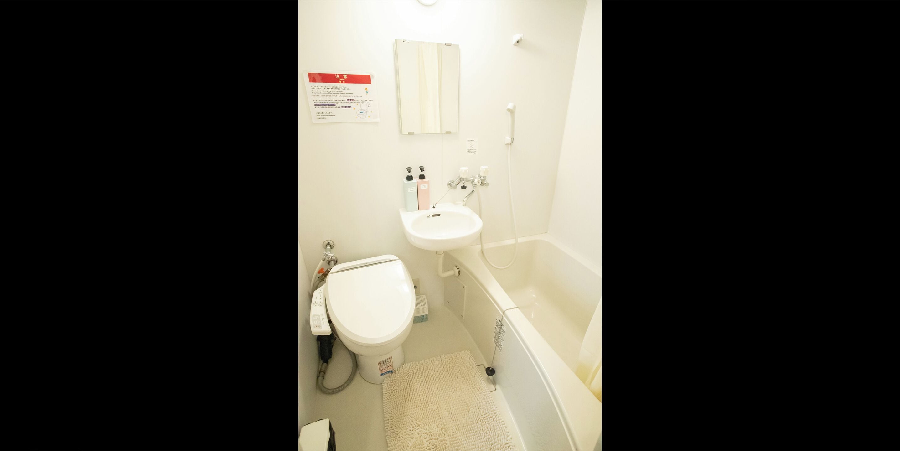 Een kleine badkamer met toilet, wastafel en bad. Er hangt een bord aan de muur en er ligt een handdoek op de vloer.