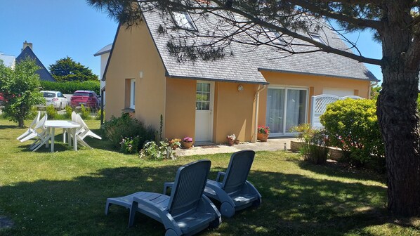 A house with a sloped roof, a front door, and a sliding glass door. There are two lounge chairs on the grass in front of the house.