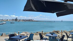 A seaside view with a dining area featuring tables and chairs, overlooking a calm sea with boats and a distant shoreline with buildings. - Bonheur de vivre au bord de l'Océan (Loctudy)