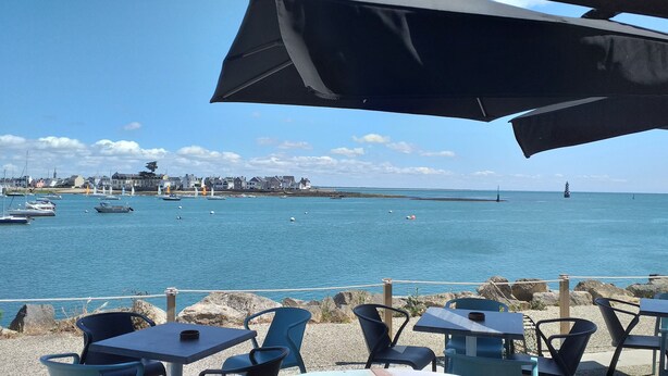 Vista al mar con un área de comedor que incluye mesas y sillas, con vista a un mar tranquilo, barcos y una costa lejana con edificios.