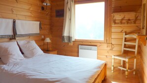 Une chambre à coucher dans une cabane en bois, avec un lit, une table de chevet, une lampe, une chaise, une fenêtre avec des rideaux et un radiateur.