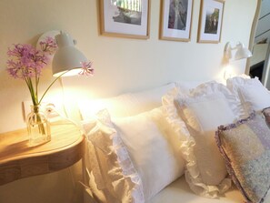 Een slaapkamer met nachtkastje, lamp, vaas met bloemen en een bed met kussens en dekbed.