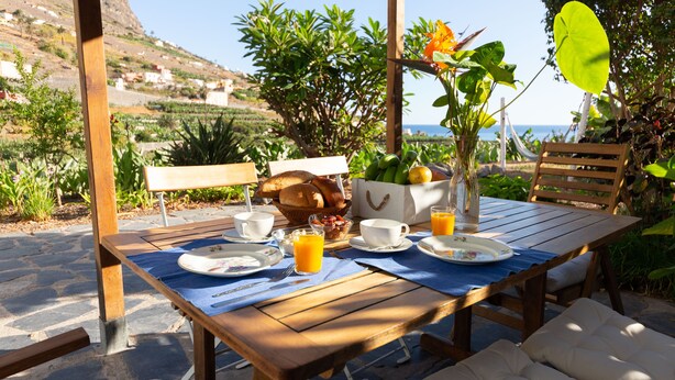 Ein Holztisch, gedeckt mit Frühstücksleckereien wie Brot, Obst und Orangensaft, in einer sonnigen Umgebung mit Blick auf das Meer und viel Grün.