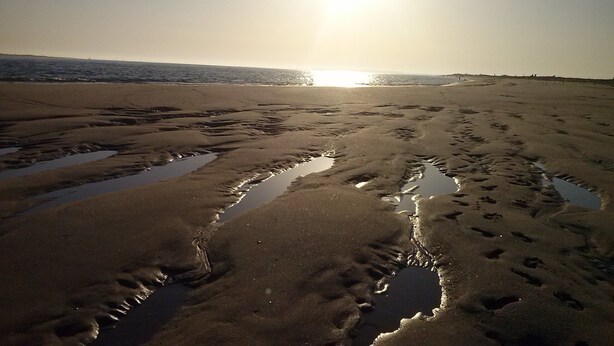 En strand med fodaftryk og vandpytter, og solen går ned i baggrunden.