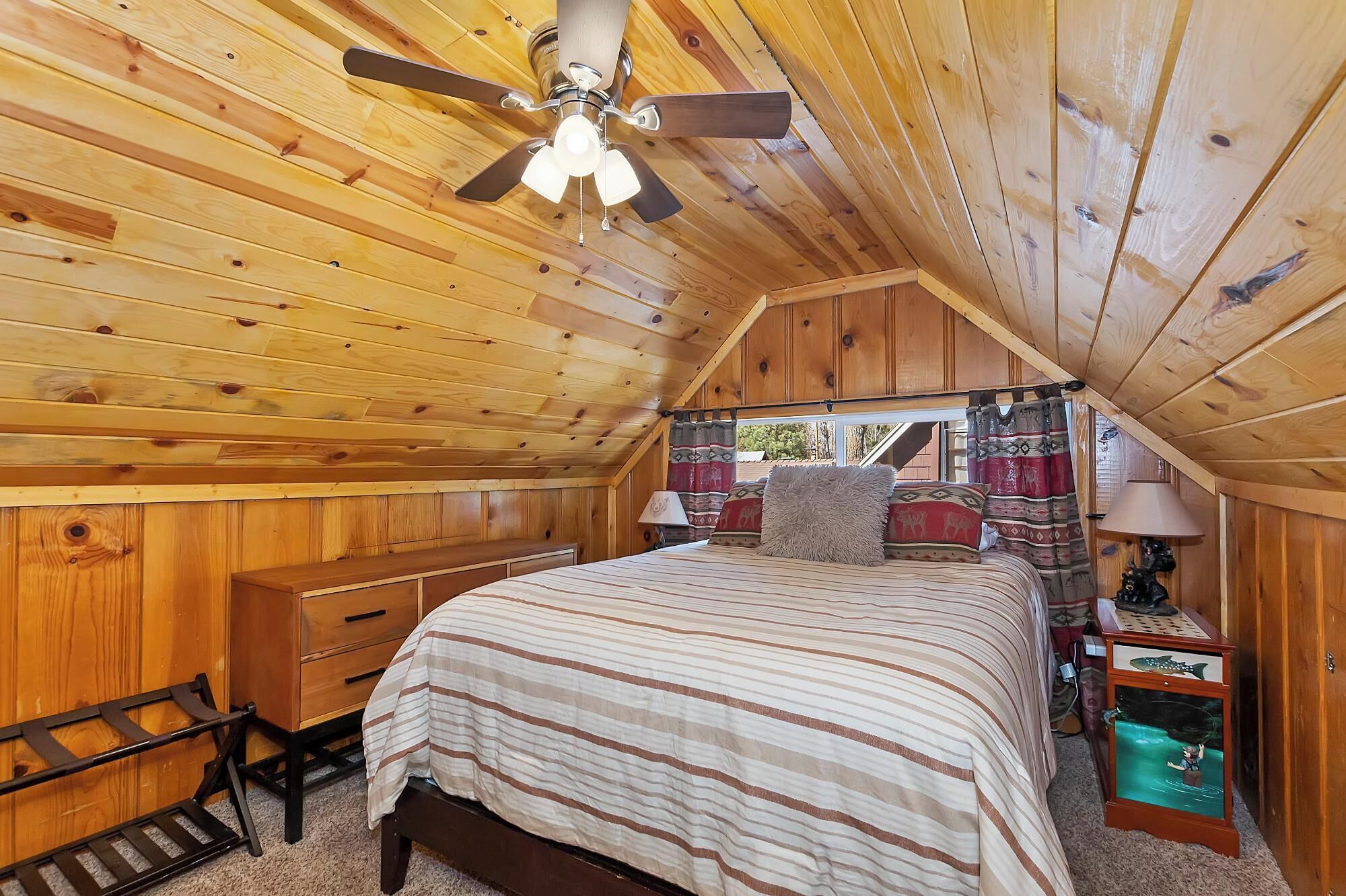 Une chambre confortable située sous les toits, avec un plafond en pente en bois, un lit avec une literie à rayures, une table de chevet avec une lampe et une petite table sur laquelle est posé un objet décoratif.