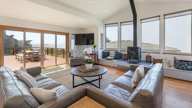 A spacious living room with a large sofa, a coffee table, and a fireplace. There is a balcony with a wooden deck and outdoor furniture. - Wonderful Views of Dillon Beach and Bodega Bay (Dillon Beach)