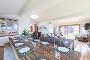 A spacious dining area with a long wooden table set for six, surrounded by modern furniture and large windows offering views of the outdoors.
