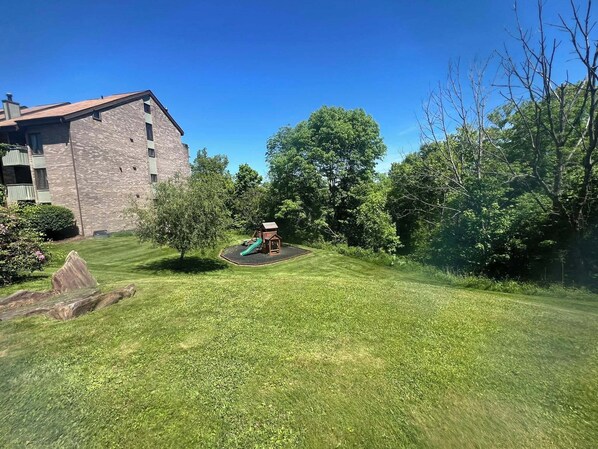 Un patio con césped, un parque infantil, árboles y un edificio al fondo.