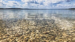 Ein klarer See mit steiniger Uferlinie und einer fernen Küste unter einem teils bewölkten Himmel.