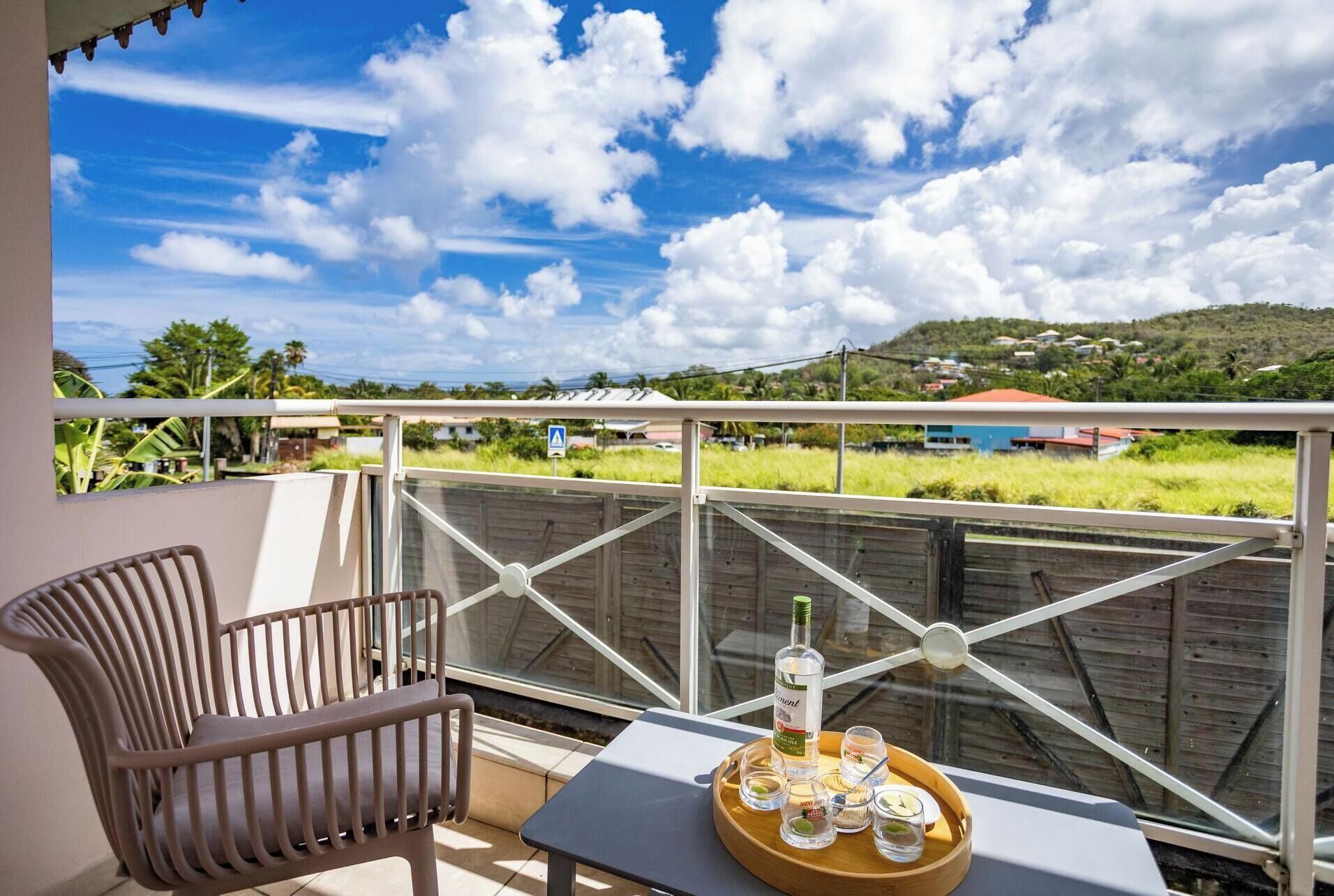A balcony with a view of a green landscape, a table with a bottle and shot glasses, and a chair.