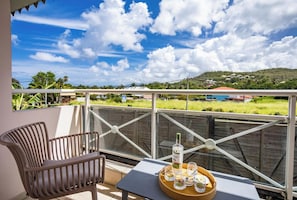 Un balcon donnant sur un paysage verdoyant, une table avec une bouteille et des verres à shooter, et une chaise.
