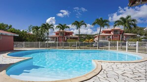 Una piscina con plataforma de piedra, rodeada por una valla blanca, y edificios con techos rojos al fondo.