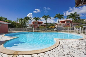 Une piscine avec une terrasse en pierre, entourée d’une clôture blanche, et des bâtiments aux toits rouges en arrière-plan.