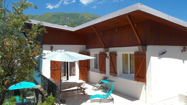 Un bâtiment moderne doté d’un toit plat, de volets en bois et d’un patio aménagé avec une table, des chaises et un parasol.