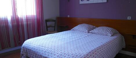 Un dormitorio con una cama, una silla, una pared con un cuadro floral y una ventana con cortinas.