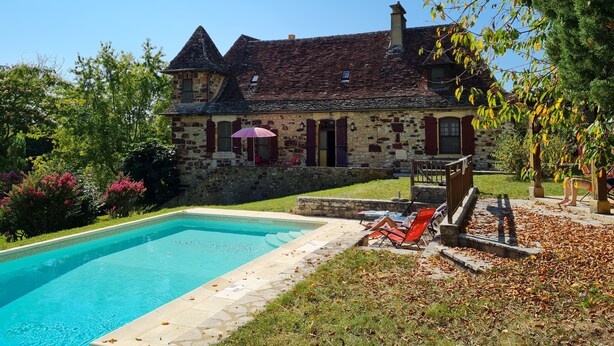 Ein Steinhaus mit Swimmingpool, roten Fensterläden und einem Garten mit Bäumen und Sträuchern.