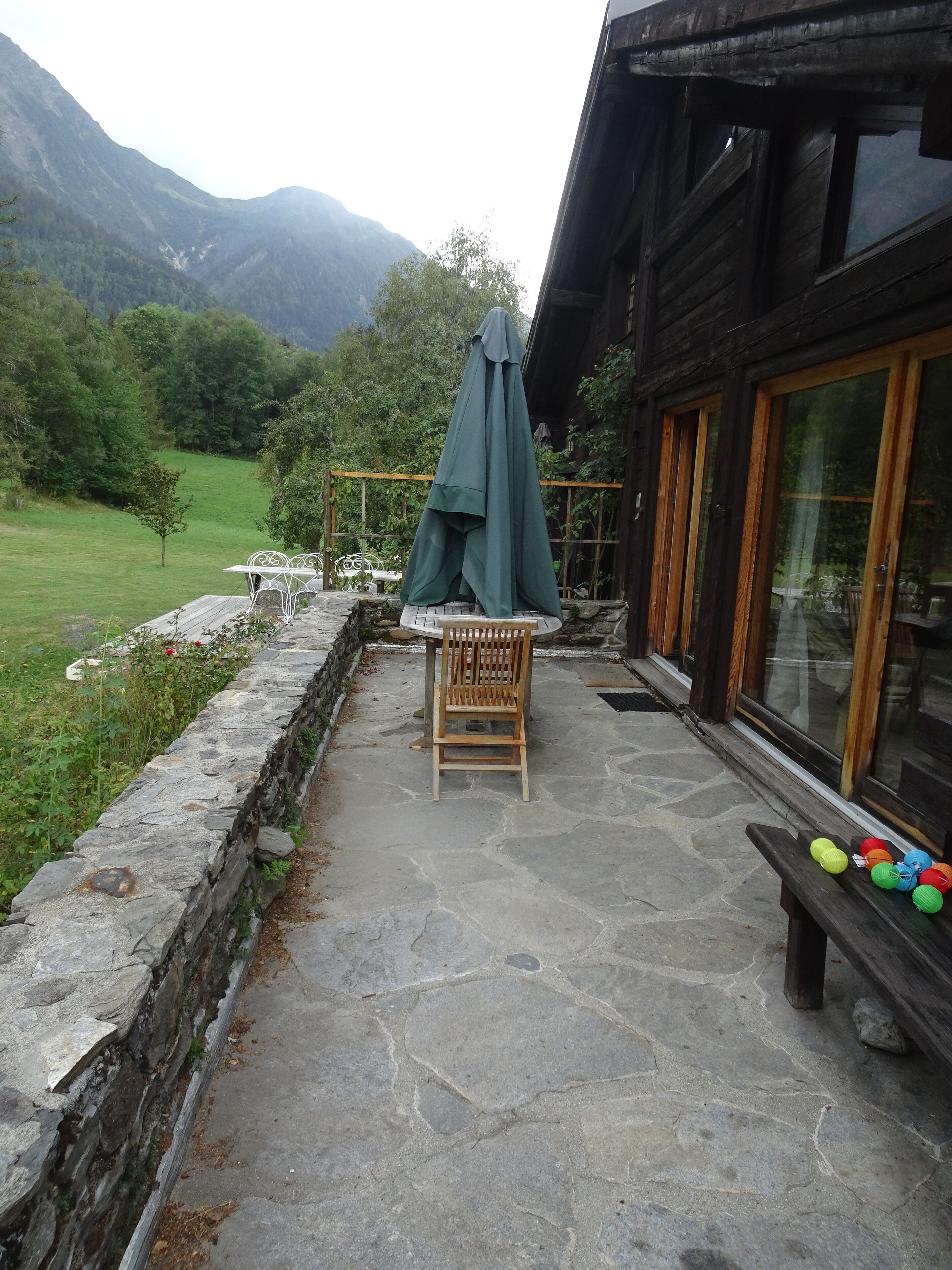 A stone patio with a wooden chair and table, a green umbrella, and colorful balls on a bench.
