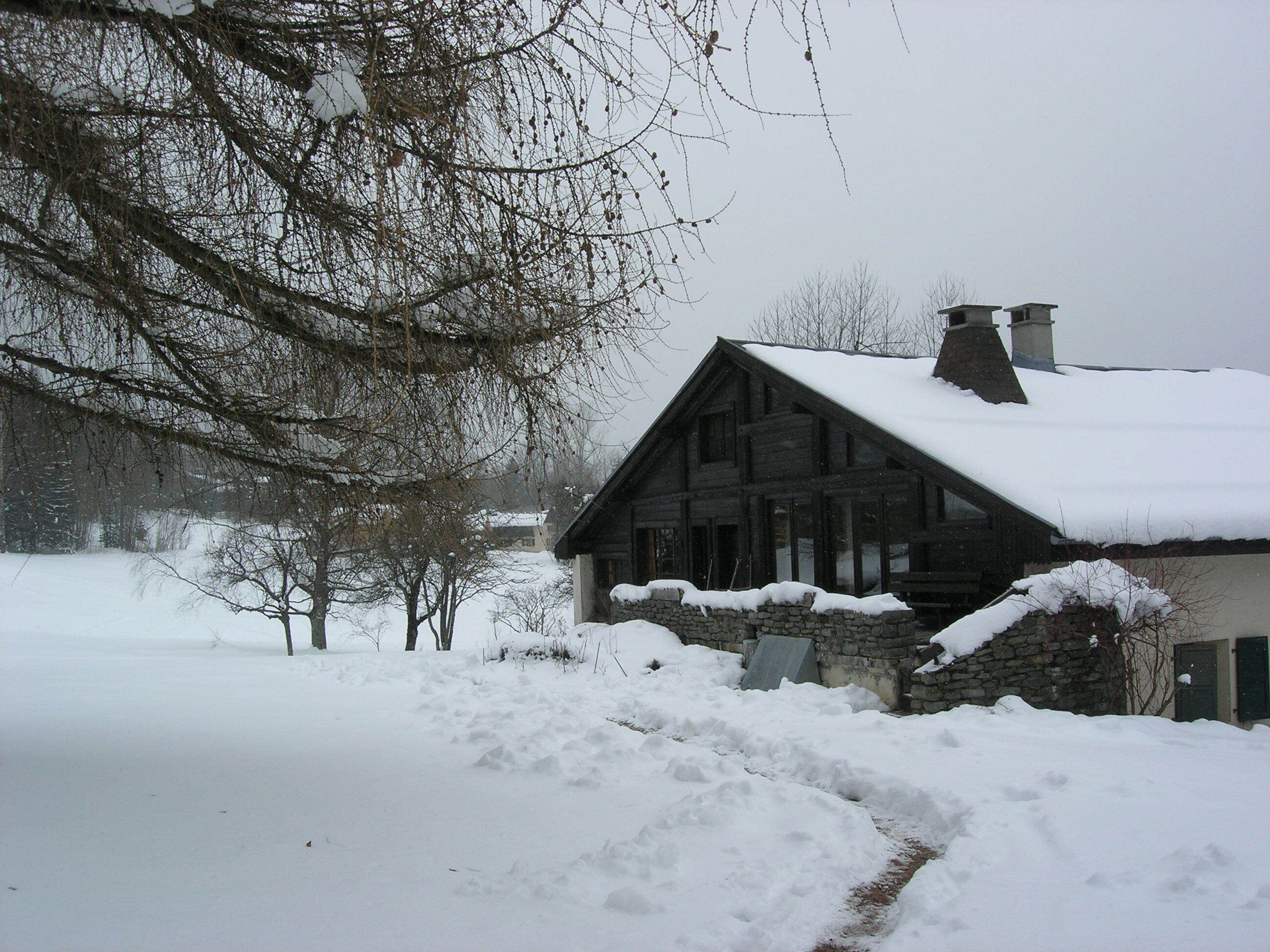 A wooden cabin with a stone foundation, surrounded by snow-covered ground and bare trees.