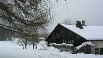 A wooden cabin with a stone foundation, surrounded by snow-covered ground and bare trees.