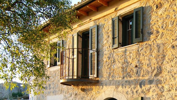 A stone building with arched windows and shutters, a balcony with a metal railing, and a tree in the foreground. - Stylishly renovated stone villa w/fantastic sea view  (Lefkas)