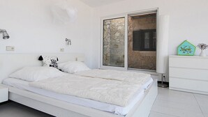A modern bedroom with a large bed, a white dresser, a window with a view of a stone wall, and a decorative plant. - Stylishly renovated stone villa w/fantastic sea view  (Lefkas)