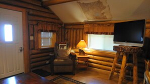 A cozy log cabin interior with a wooden dining table, a television on a wooden stand, a chair with a bear-themed pillow, and a window with blinds. - WHISKEY SPRINGS CABINS #1
Cozy and delightful, in town convenience (West Yellowstone)