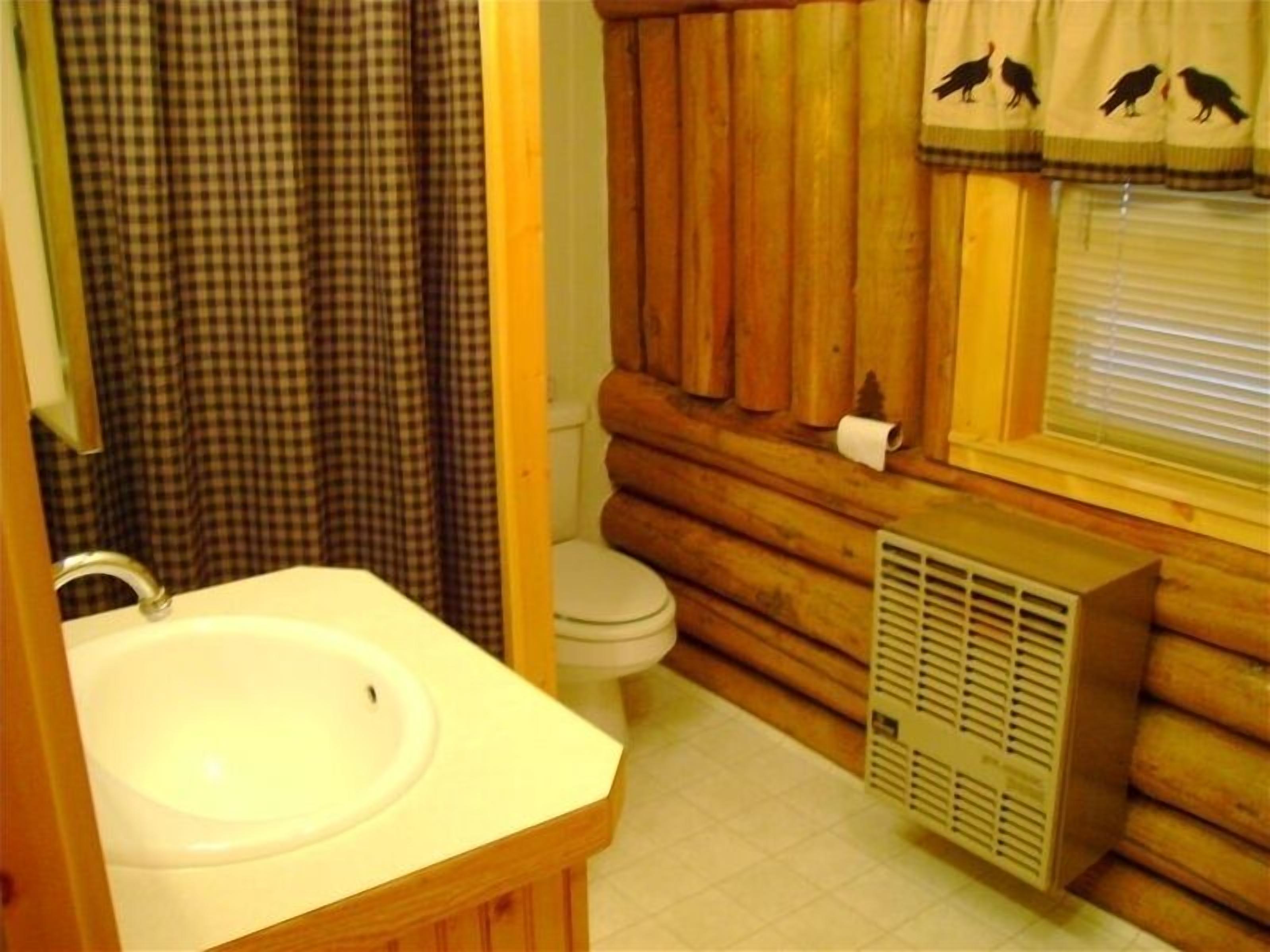A bathroom with a wooden log wall, a white sink, a toilet, and a window with a curtain featuring bird silhouettes.