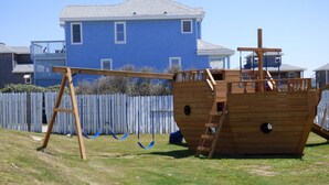 Eine hölzerne Piratenburg-Spielanlage in einem Garten mit weißem Zaun und Häusern im Hintergrund.